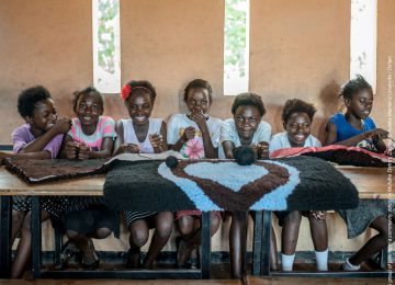 Mädchen in Sambia sitzen auf einem Tisch und lachen, vor Ihnen ein gewebtes Herz