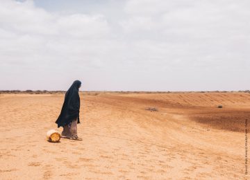 eine Frau mit einem leeren Wasserkanister steht in Afrika vor einem ausgetrockneten Fluss. Um sie herum nur Wüste.