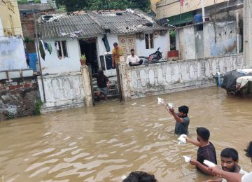 Das Foto zeigt eine überschwemmte Straße in Indien. Männer stehen im Wasser und überreichen Lebensmittel an einen Haushalt der stark betroffen ist von den Überflutungen.